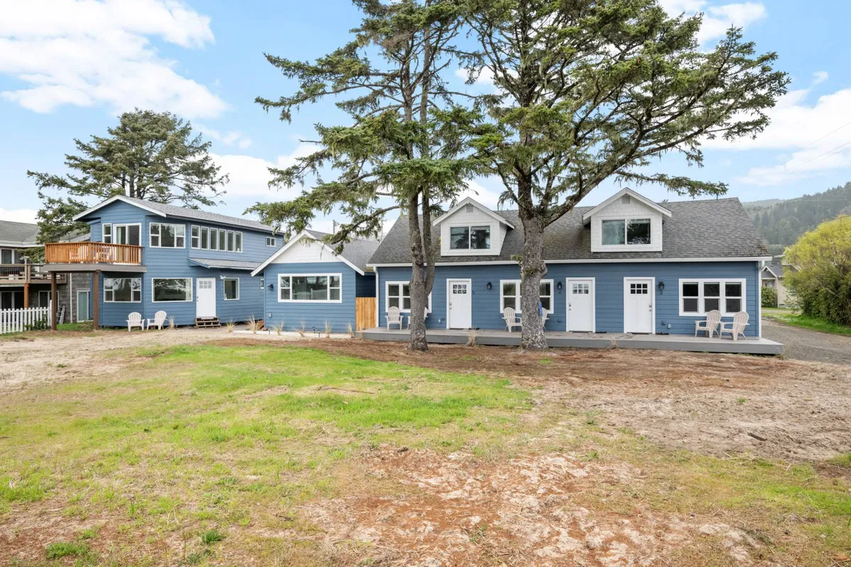 A friendly exterior shot showing the blue siding of the house with large windows, framed by trees and featuring a well-appointed outdoor seating area. The setting is ideal for relaxation, family gatherings, and informal socializing near nature.