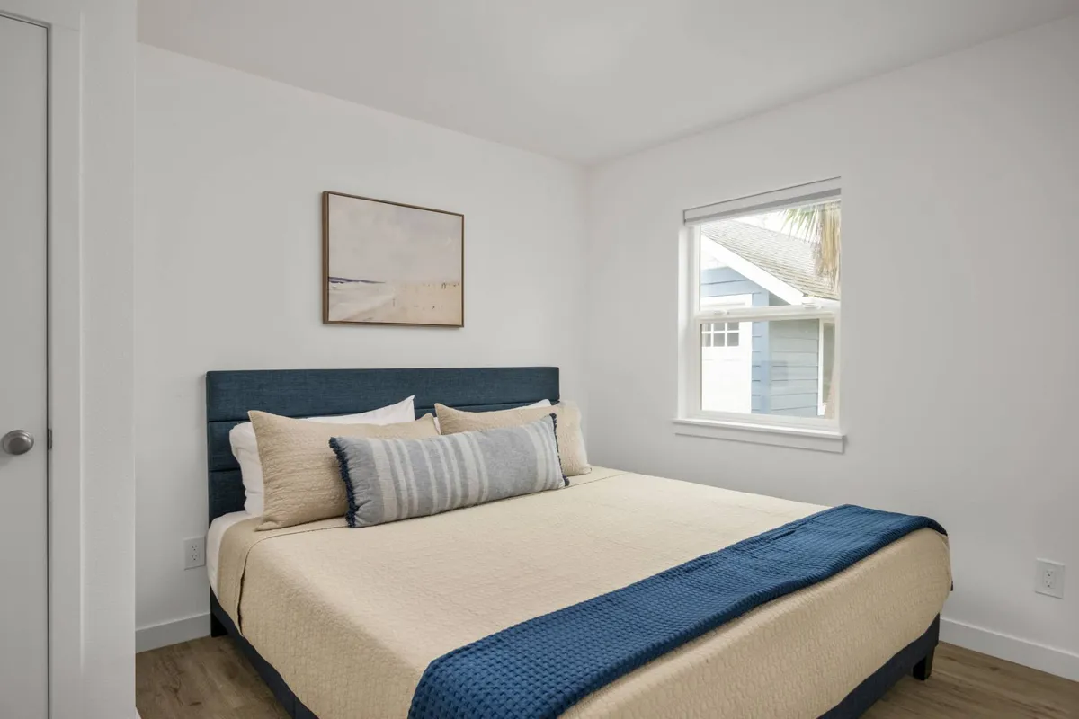 The focus is on a serene bedroom with a plush bed dressed in blue accents, decorative pillows, and artwork on the wall. Sunlight filters through a window, casting a calming glow across the crisp linens.