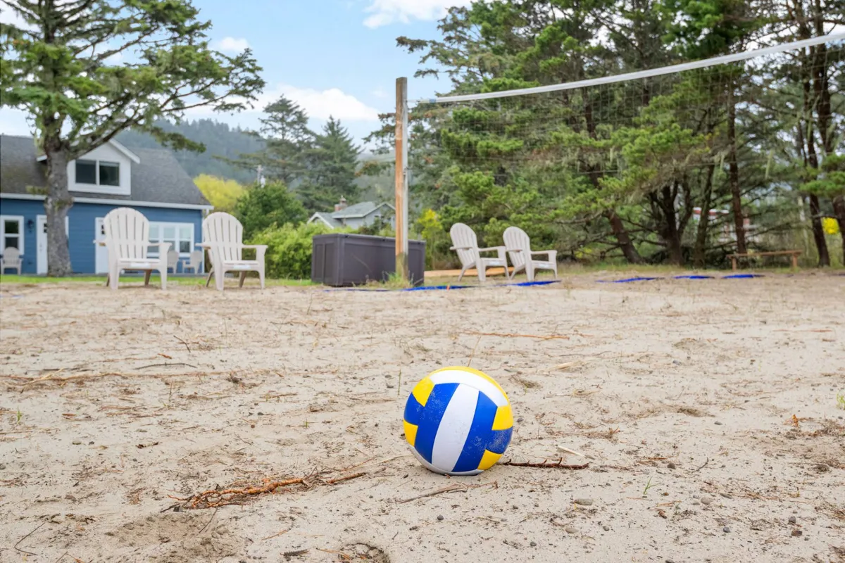 This image captures a volleyball net set up on a sandy outdoor area with Adirondack chairs nearby, a house in the background, and trees surrounding the space. The scene conveys active, family-friendly fun right on the property, with space for both energetic play and relaxed spectating.