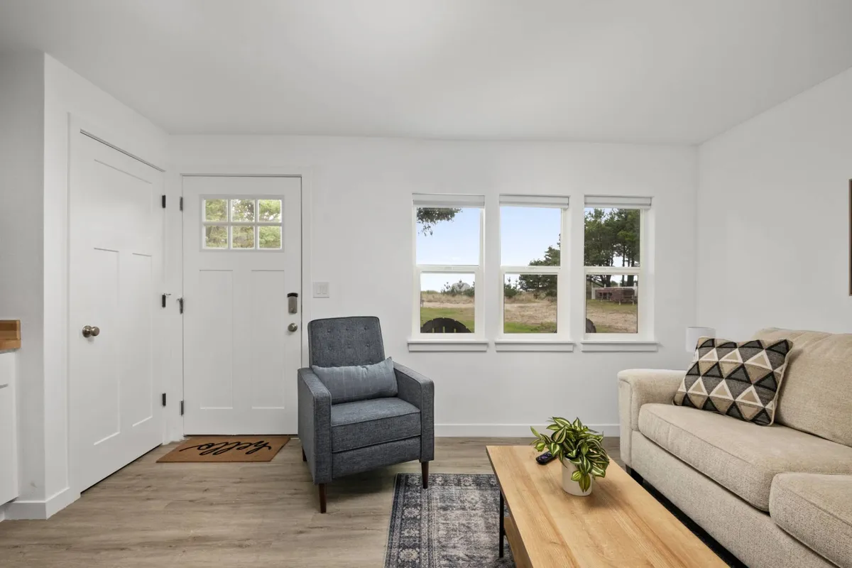 A cozy, sunlit living area with a welcoming front door, large windows, an inviting armchair, plush sofa, and a coffee table. The space is arranged to foster relaxation, reading, and socializing, creating a calm and homey atmosphere.