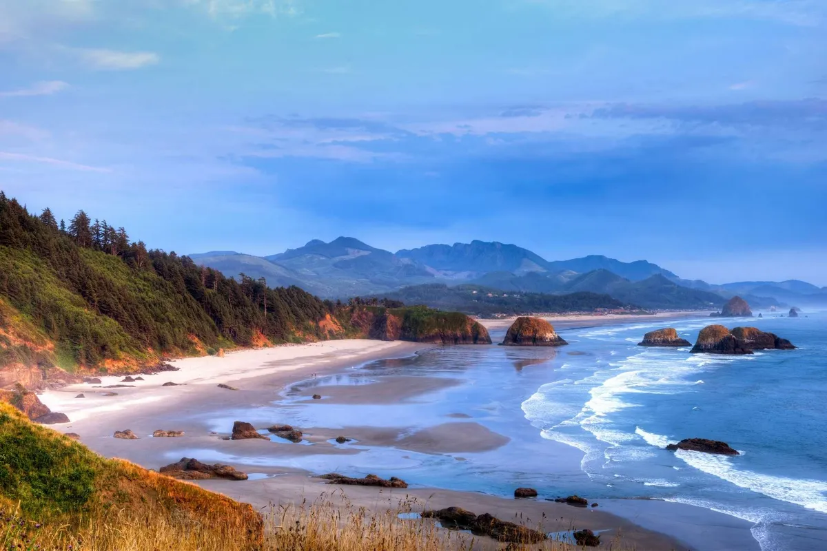 A sweeping view of Rockaway Beach featuring golden sand, the glistening ocean, dramatic rock formations, and distant mountains beneath a tranquil sky. The scene captures the natural beauty and openness of the beach, inviting walks and romantic photography, especially with the Twin Rocks Arch visible.