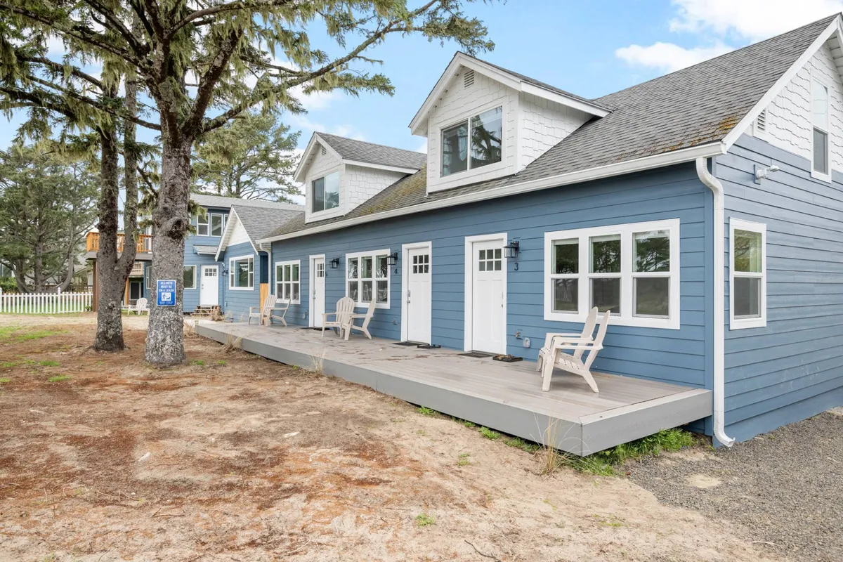 The blue exterior of one of the houses is highlighted alongside inviting porch seating, tall trees, and a serene, natural setting. The space is ideal for relaxing after group activities, enjoying sunset views, and casual socializing outdoors.