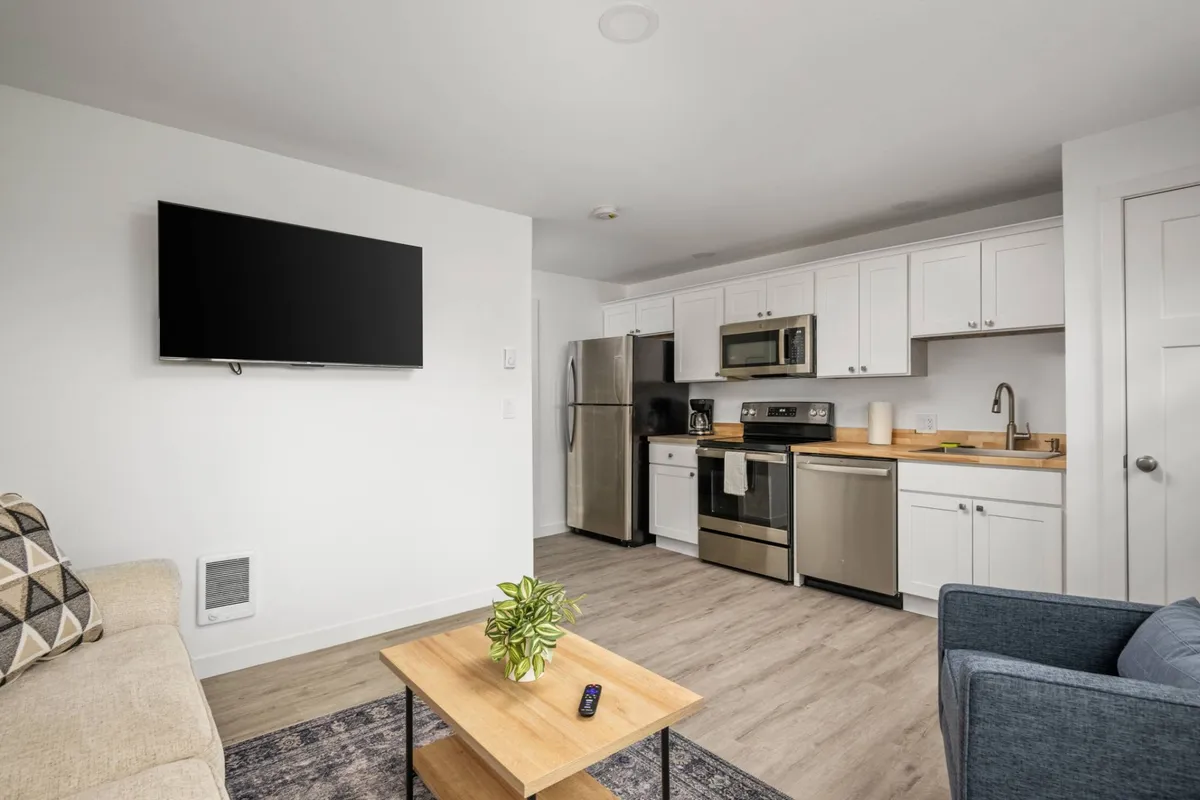 A cozy and contemporary open-concept space featuring a plush sofa, coffee table, and a TV, with the kitchen area and its modern appliances visible in the background. The setup invites guests to enjoy both relaxation and culinary experiences, with clean lines and stylish décor throughout.