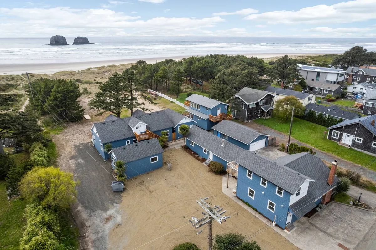 A stunning exterior beachfront view featuring blue houses nestled among trees, with direct access to sandy shores and picturesque rock formations in the background. The open setting invites beach walks and outdoor play.