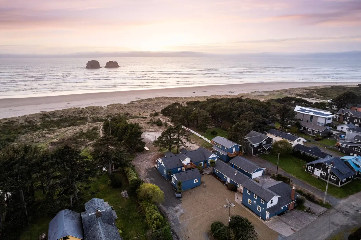 The image showcases the condo's beachfront location with expansive ocean views, iconic Twin Rocks formations rising just offshore, a sandy stretch perfect for play, and surrounding coastal houses and trees that create a sense of tranquil retreat. The scene suggests serene sunsets and fun-filled days by the sea.