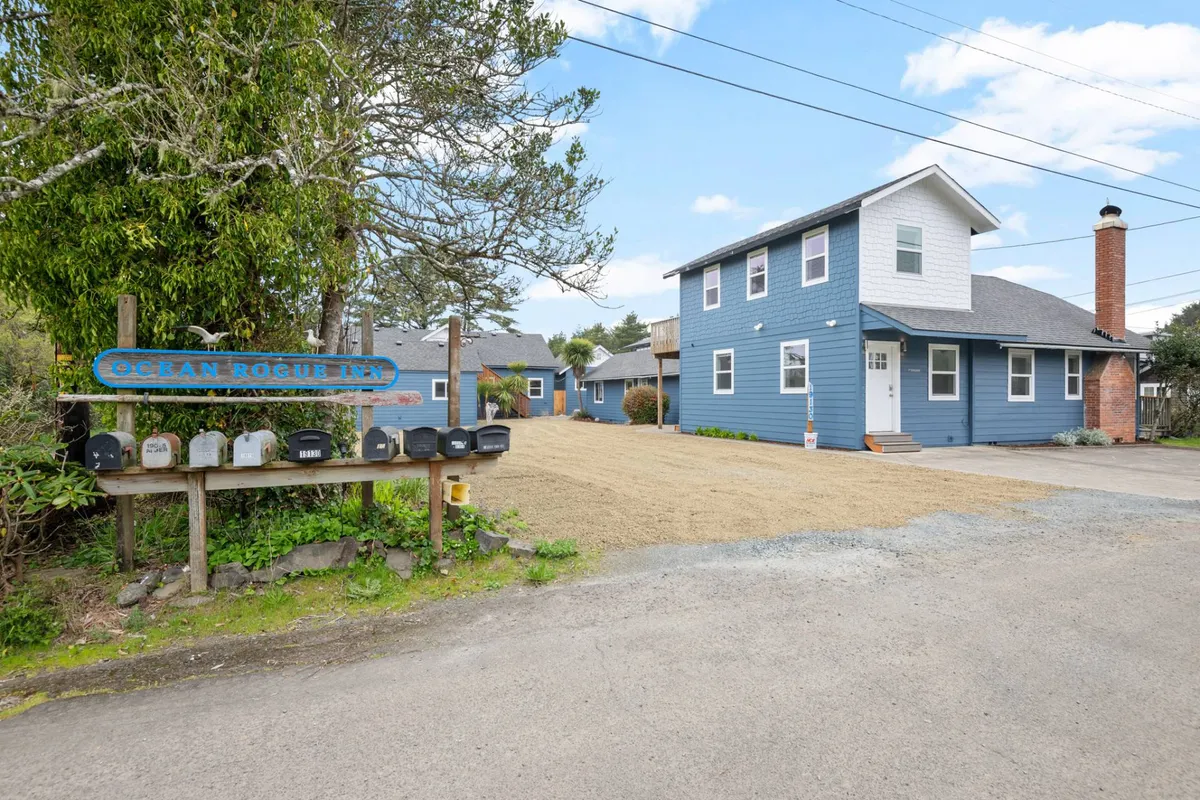The exterior of Ocean Rogue Inn is shown with its distinctive blue facade, a welcoming sign, and a mailbox setup. The scene includes pathways and garden elements, suggesting easy access to surrounding areas for walks and exploration.