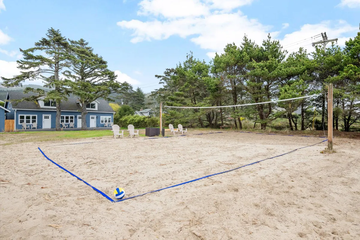 A lively exterior scene focused on the sandy volleyball court, bordered by beach chairs and surrounded by trees. The court is set up for play, inviting guests to join in a spirited match or casual social gathering as the sun sets.