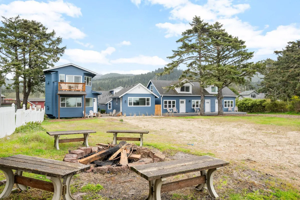 This image captures the inviting fire-pit area surrounded by blue houses and a large tree. People are gathered around the fire, fostering a sense of community and warmth. The relaxed atmosphere is enhanced by the open space and the presence of outdoor games and barbecue activities.